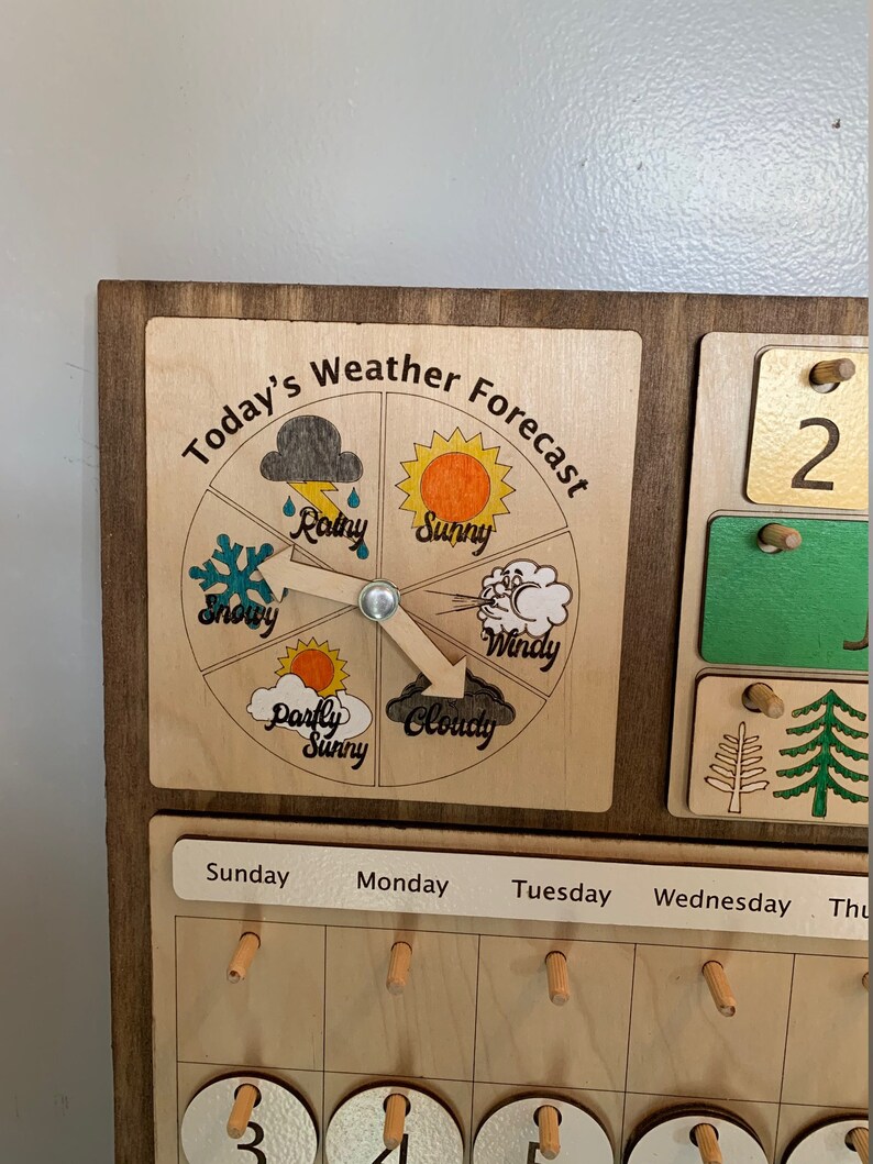 Pode incluir: Um quadro de previs&atilde;o do tempo de madeira com um dial rotativo que apresenta diferentes &iacute;cones meteorol&oacute;gicos: ensolarado, nublado, chuvoso, nevado, ventoso e parcialmente ensolarado. O quadro tamb&eacute;m inclui um calend&aacute;rio com os dias da semana e os dias numerados.