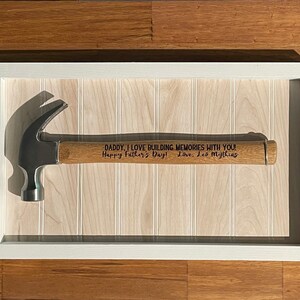 May include: A framed hammer with a wooden handle. The handle is engraved with the message "DADDY, I LOVE BUILDING MEMORIES WITH YOU! Happy Father's Day! Love, Les Mythins".