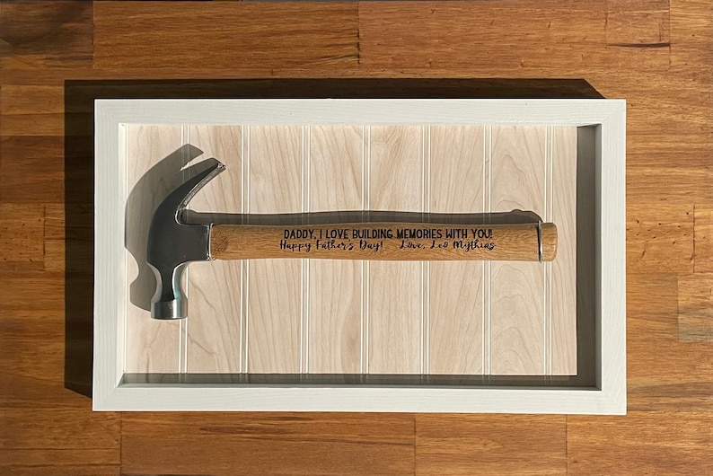 May include: A framed hammer with a wooden handle. The handle is engraved with the message "DADDY, I LOVE BUILDING MEMORIES WITH YOU! Happy Father's Day! Love, Les Mythins".