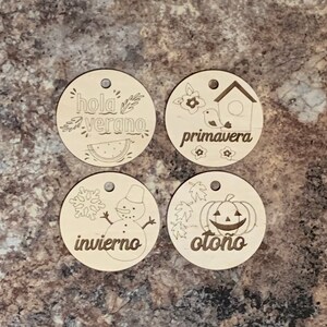May include: Four wooden ornaments with different seasonal themes. The top left ornament features a watermelon and the text "hola verano". The top right ornament features a birdhouse and the text "primavera". The bottom left ornament features a snowman and the text "invierno". The bottom right ornament features a pumpkin and the text "oto&ntilde;o".