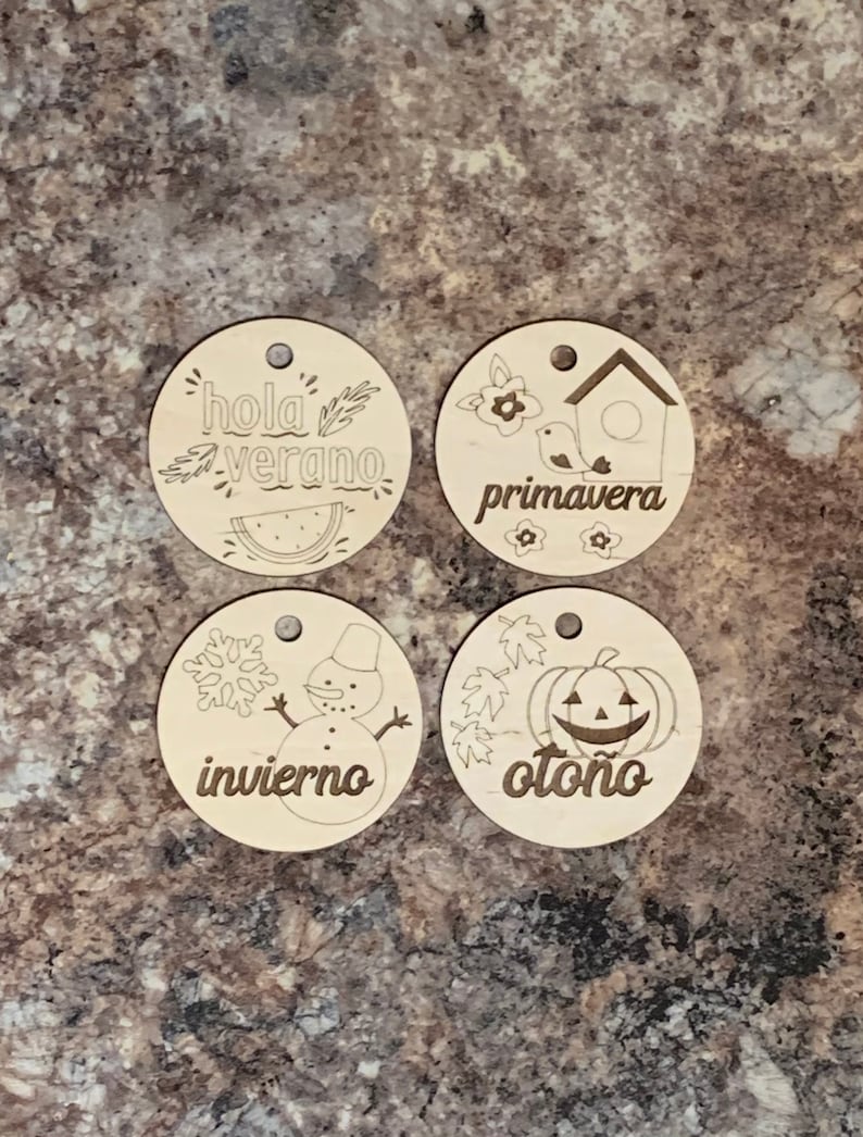 May include: Four wooden ornaments with different seasonal themes. The top left ornament features a watermelon and the text "hola verano". The top right ornament features a birdhouse and the text "primavera". The bottom left ornament features a snowman and the text "invierno". The bottom right ornament features a pumpkin and the text "oto&ntilde;o".