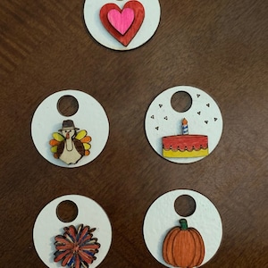 Pode incluir: Cinco ornamentos redondos de madeira com um furo no topo. Cada ornamento tem um design diferente: um cora&ccedil;&atilde;o rosa, um peru, um bolo de anivers&aacute;rio, uma flor e uma ab&oacute;bora.