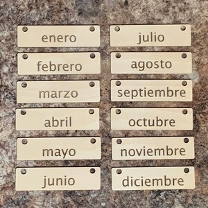 May include: Twelve wooden tags with the names of the months of the year in Spanish. The tags are light brown and have two holes at the top.