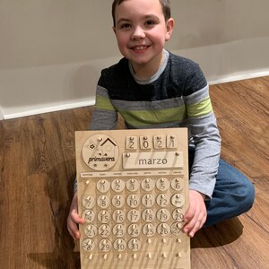 May include: A wooden calendar with a birdhouse design and the word "primavera" on it. The calendar is for March 2021 and has a movable piece for each day of the month. The calendar is made of light brown wood and has a natural finish.
