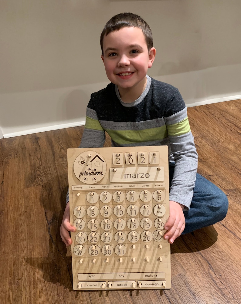 May include: A wooden calendar with a birdhouse design and the word "primavera" on it. The calendar is for March 2021 and has a movable piece for each day of the month. The calendar is made of light brown wood and has a natural finish.