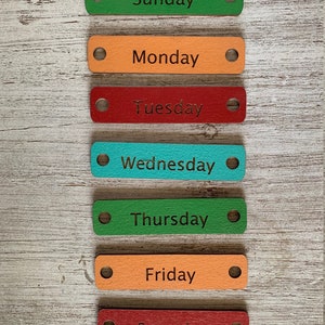 Pode incluir: Sete etiquetas de madeira com os dias da semana escritos nelas. As etiquetas s&atilde;o pintadas em cores diferentes: verde, laranja, vermelho, turquesa e marrom. Cada etiqueta tem um furo perfurado na parte superior.