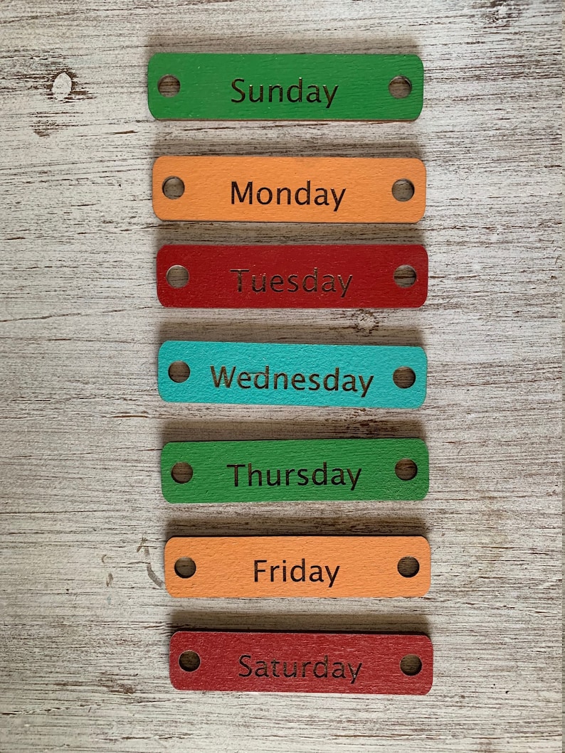 Pode incluir: Sete etiquetas de madeira com os dias da semana escritos nelas. As etiquetas s&atilde;o pintadas em cores diferentes: verde, laranja, vermelho, turquesa e marrom. Cada etiqueta tem um furo perfurado na parte superior.