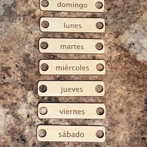 May include: Seven wooden labels with rounded edges and holes at each end. The labels are labeled with the days of the week in Spanish: Domingo, Lunes, Martes, Mi&eacute;rcoles, Jueves, Viernes, S&aacute;bado.