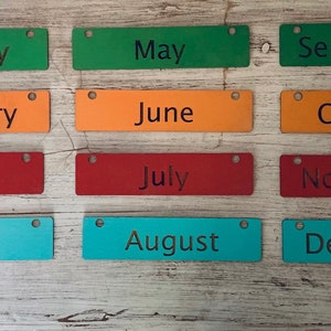 Pode incluir: Doze etiquetas retangulares de madeira com os meses do ano impressos nelas. As etiquetas s&atilde;o de diferentes cores: verde, laranja, vermelho, azul e turquesa.