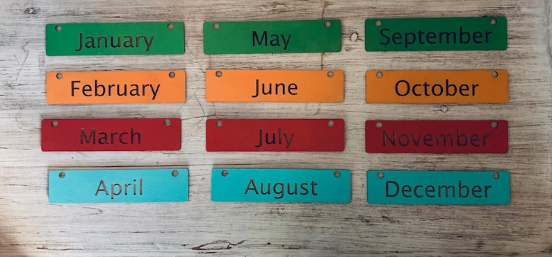 Pode incluir: Doze etiquetas retangulares de madeira com os meses do ano impressos nelas. As etiquetas s&atilde;o de diferentes cores: verde, laranja, vermelho, azul e turquesa.
