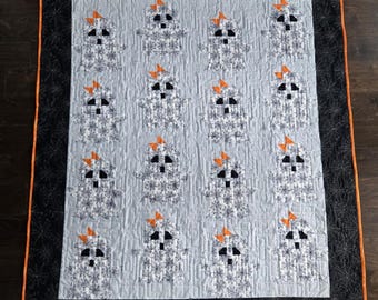 Könnte beinhalten: Eine handgefertigte Steppdecke mit einem sich wiederholenden Muster von Geisterfiguren mit orangefarbenen Schleifen. Die Geister sind weiß mit schwarzen Augen und Mündern, vor einem hellblauen Hintergrund. Die Steppdecke hat einen schwarzen Rand mit orangefarbener Einfassung und Spinnennetz-Design.