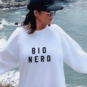 May include: White sweatshirt with black text that reads "BIO NERD". The sweatshirt is worn by a person standing on a rocky beach.