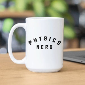 May include: White ceramic coffee mug with a black inscription that reads "PHYSICS NERD". The mug is sitting on a wooden table next to a laptop.