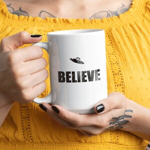 K&ouml;nnte beinhalten: Eine wei&szlig;e Keramiktasse mit schwarzem Henkel und dem Wort "BELIEVE" aufgedruckt. Oberhalb des Wortes "BELIEVE" befindet sich ein kleines Schwarzwei&szlig;bild einer fliegenden Untertasse.