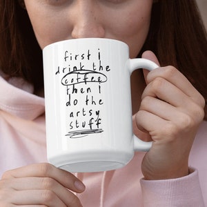 May include: White ceramic mug with black text that reads "first I drink the coffee then I do the artsy stuff". The text is handwritten and circled. A person is holding the mug.
