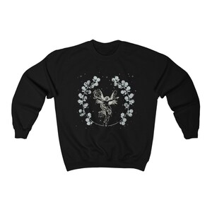May include: Black sweatshirt with a white graphic of a fairy with wings spread, surrounded by a wreath of flowers and stars.