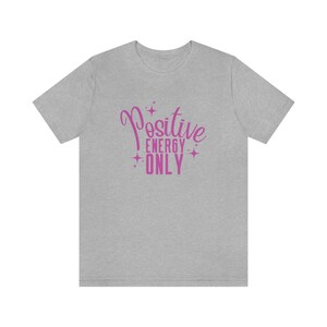 Op de afbeelding: Een grijs t-shirt met een roze afbeelding met de tekst "Positive Energy Only" met sterren rond de tekst.