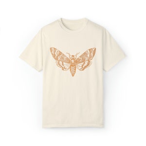 May include: A cream colored t-shirt with a vintage illustration of a moth in orange on the front.