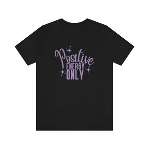 Op de afbeelding: Zwart T-shirt met een paars ontwerp met de tekst "Positive Energy Only" met sterren rond de tekst.