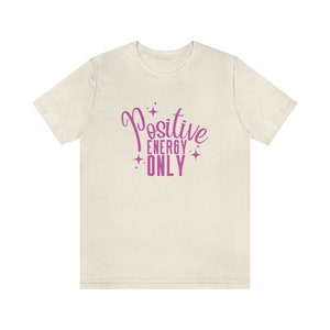 Op de afbeelding: Een wit T-shirt met een roze afbeelding met de tekst "Positive Energy Only" met sterren rond de tekst.