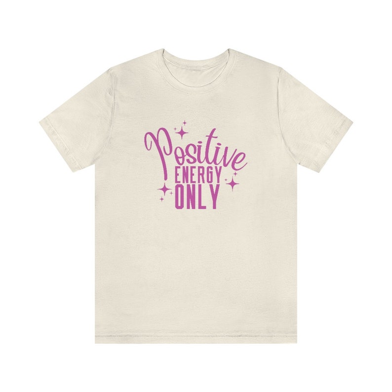 Op de afbeelding: Een wit T-shirt met een roze afbeelding met de tekst "Positive Energy Only" met sterren rond de tekst.