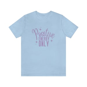 Op de afbeelding: Lichtblauw T-shirt met een paarse grafische afbeelding met de tekst "Positive Energy Only" met sterren rond de tekst.