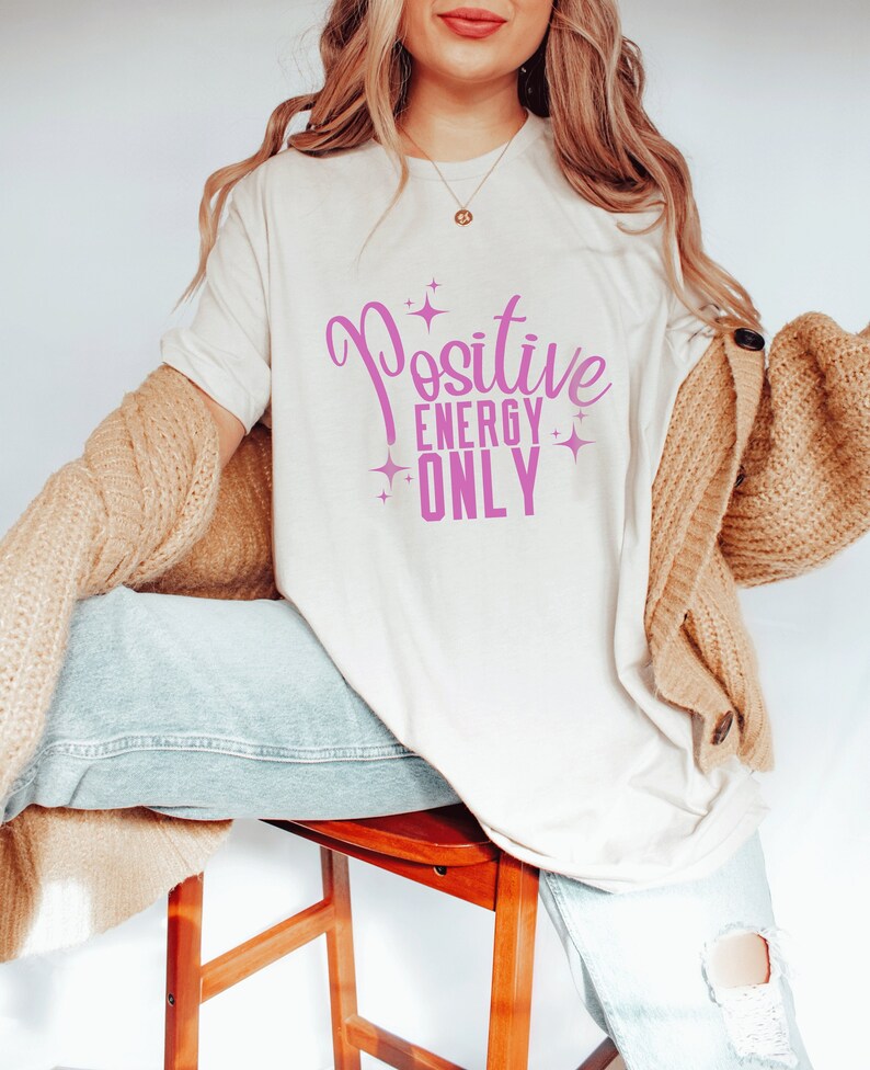 Op de afbeelding: Een wit T-shirt met de tekst "Positive Energy Only" in roze letters met sterren rond de tekst.