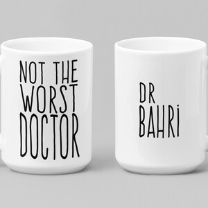 May include: White ceramic mug with black text. One side reads "NOT THE WORST DOCTOR" and the other side says "DR BAHRI". The mug has a large handle and is suitable for hot or cold beverages.