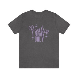 Op de afbeelding: Een donker grijs t-shirt met de tekst "Positive Energy Only" in paarse letters met glinsterende sterren.