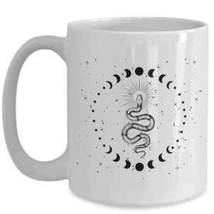 Pode incluir: Caneca de caf&eacute; de cer&acirc;mica branca com uma ilustra&ccedil;&atilde;o em preto e branco de uma cobra com uma coroa na cabe&ccedil;a, cercada por um c&iacute;rculo de luas crescentes e estrelas.