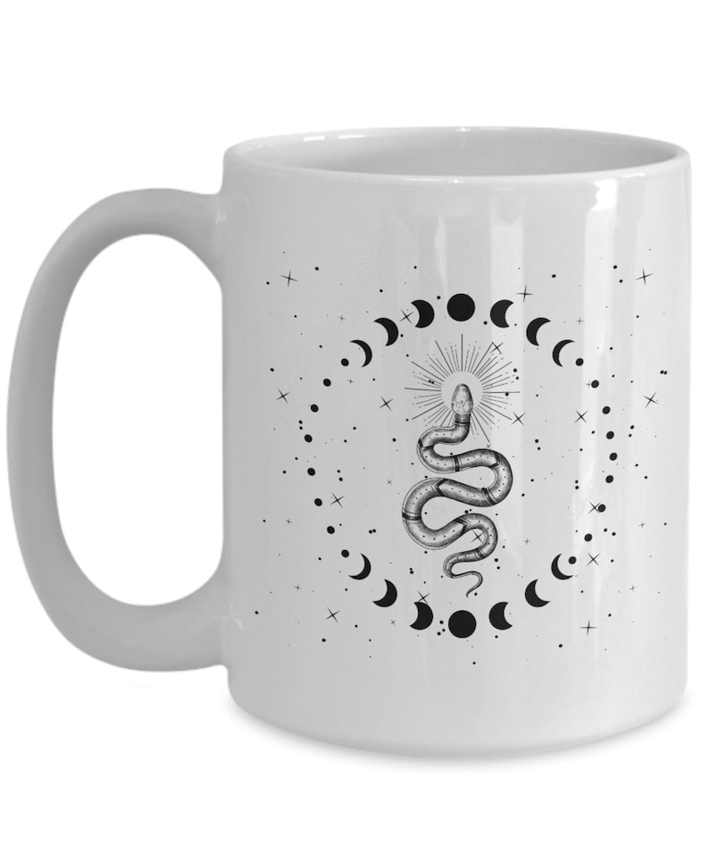Pode incluir: Caneca de caf&eacute; de cer&acirc;mica branca com uma ilustra&ccedil;&atilde;o em preto e branco de uma cobra com uma coroa na cabe&ccedil;a, cercada por um c&iacute;rculo de luas crescentes e estrelas.