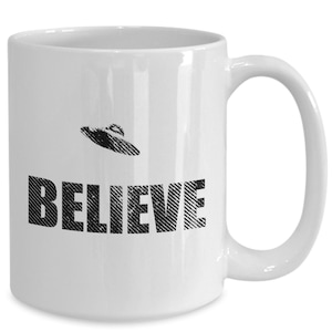 K&ouml;nnte beinhalten: Wei&szlig;e Keramik-Kaffeetasse mit einer schwarz-wei&szlig;en Grafik eines UFOs und dem Wort "BELIEVE" darauf.