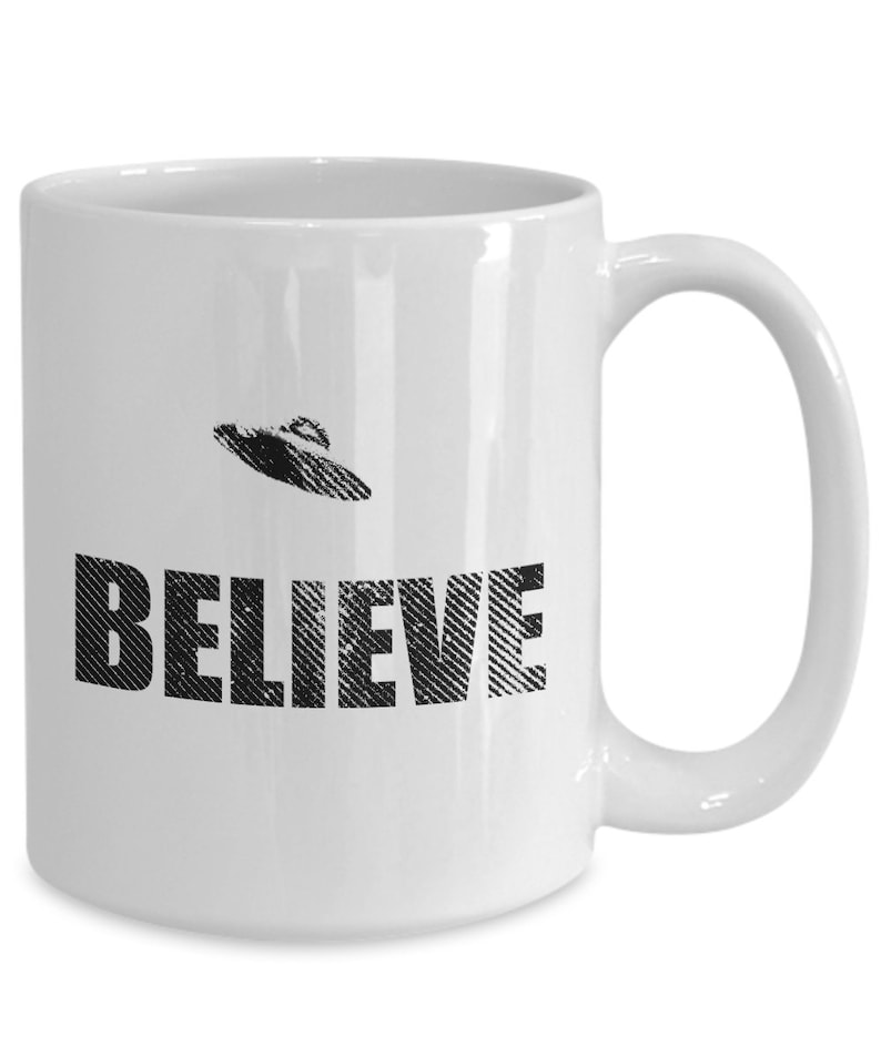 K&ouml;nnte beinhalten: Wei&szlig;e Keramik-Kaffeetasse mit einer schwarz-wei&szlig;en Grafik eines UFOs und dem Wort "BELIEVE" darauf.