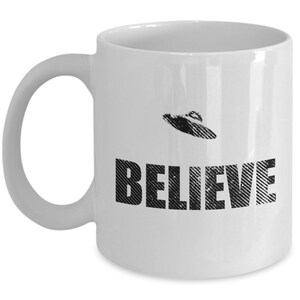 K&ouml;nnte beinhalten: Wei&szlig;e Keramik-Kaffeetasse mit einem Schwarzwei&szlig;bild eines UFOs und dem Wort "BELIEVE" darauf gedruckt.
