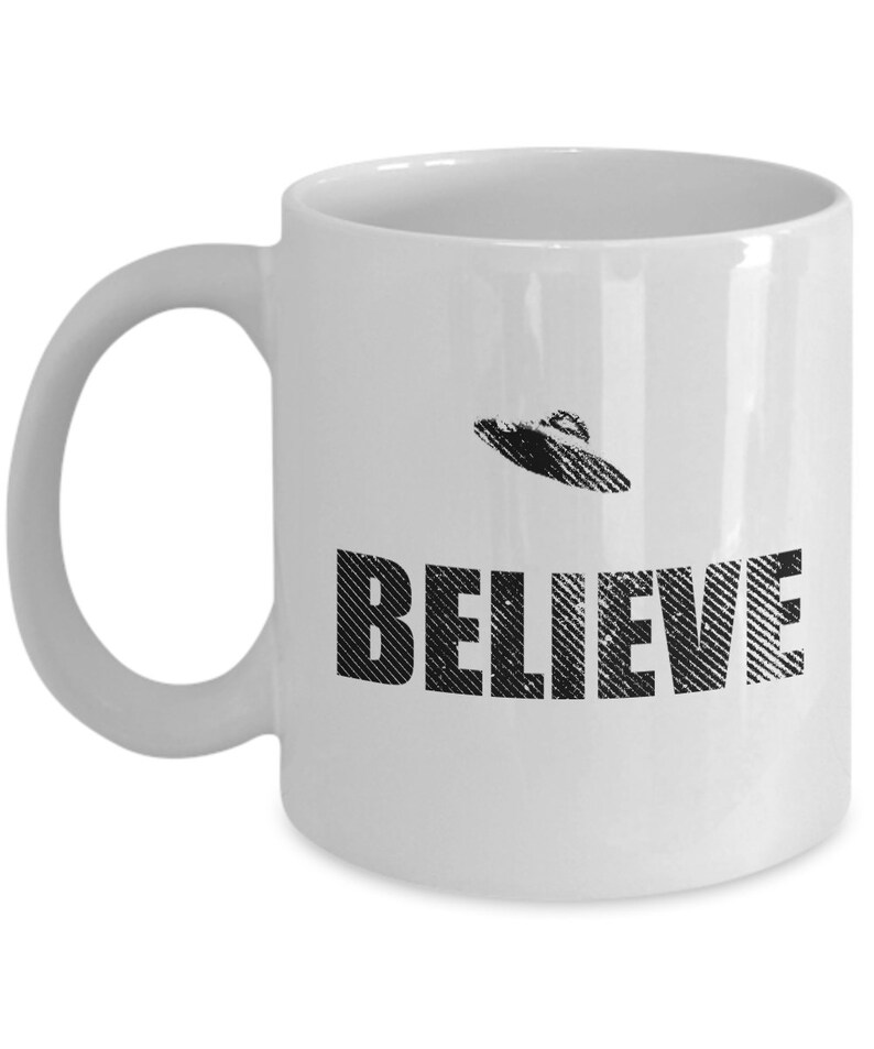 K&ouml;nnte beinhalten: Wei&szlig;e Keramik-Kaffeetasse mit einem Schwarzwei&szlig;bild eines UFOs und dem Wort "BELIEVE" darauf gedruckt.