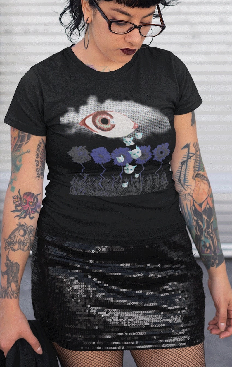 May include: Black t-shirt with a graphic of a large eye with a tear falling from it. The tear is made up of small white cats. The eye is surrounded by a cloud and blue flowers with white cats on them.