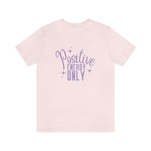 Op de afbeelding: Een roze T-shirt met de tekst "Positive Energy Only" in paarse letters met sterren rond de tekst.