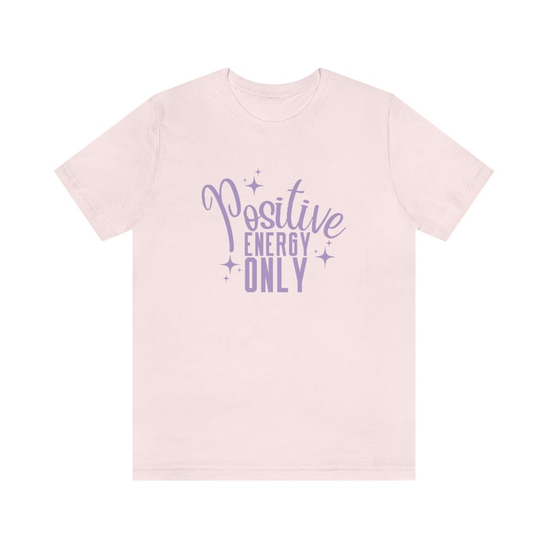 Op de afbeelding: Een roze T-shirt met de tekst "Positive Energy Only" in paarse letters met sterren rond de tekst.