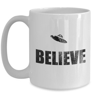 K&ouml;nnte beinhalten: Wei&szlig;e Keramik-Kaffeetasse mit einem Schwarzwei&szlig;bild eines UFOs und dem Wort "BELIEVE" darauf gedruckt.