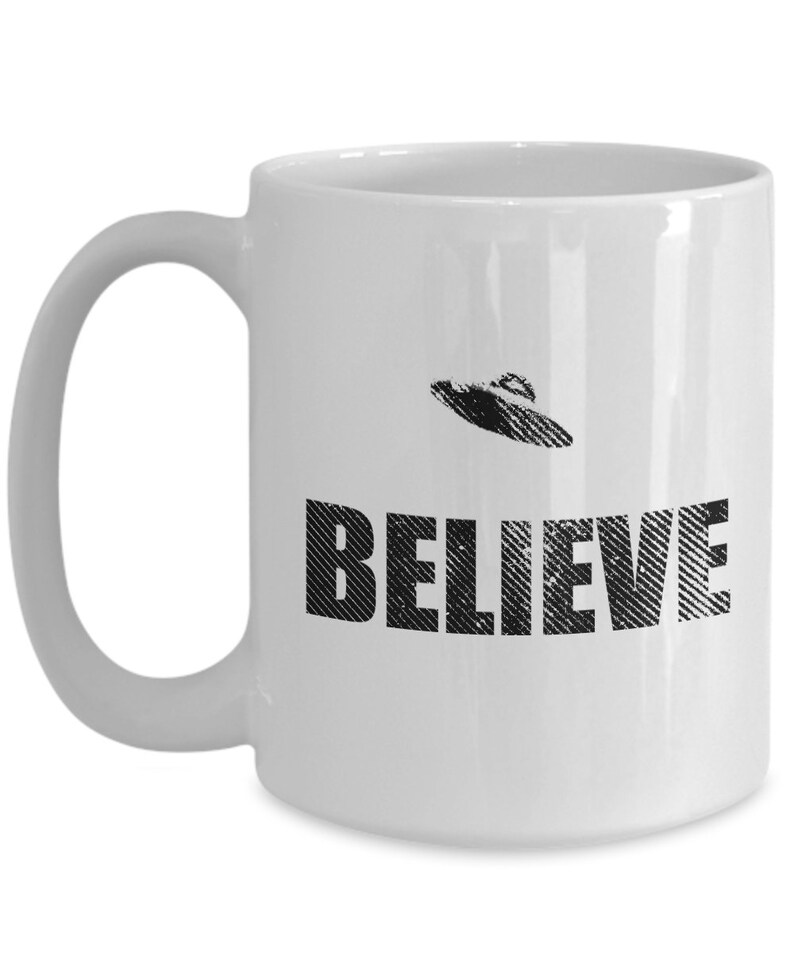 K&ouml;nnte beinhalten: Wei&szlig;e Keramik-Kaffeetasse mit einem Schwarzwei&szlig;bild eines UFOs und dem Wort "BELIEVE" darauf gedruckt.