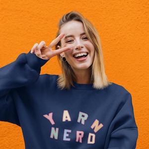May include: A navy blue sweatshirt with the words "YARN NERD" in pastel colors. The person in the image is smiling and making a peace sign. The background is a solid orange color.