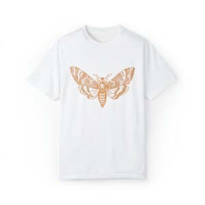 May include: White t-shirt with a brown outline of a moth printed on the front.