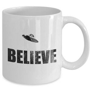 K&ouml;nnte beinhalten: Wei&szlig;e Keramik-Kaffeetasse mit einer schwarz-wei&szlig;en Grafik eines UFOs und dem Wort "BELIEVE" darauf.