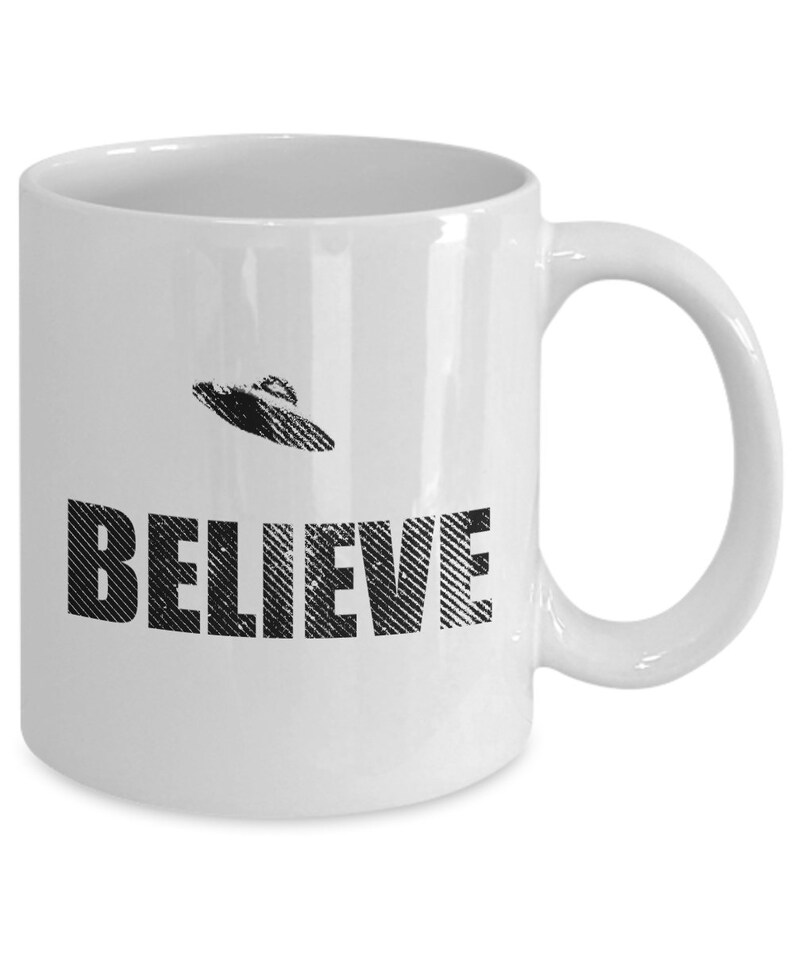 K&ouml;nnte beinhalten: Wei&szlig;e Keramik-Kaffeetasse mit einer schwarz-wei&szlig;en Grafik eines UFOs und dem Wort "BELIEVE" darauf.