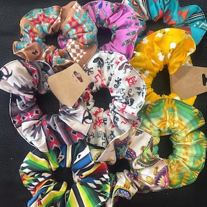 May include: A collection of colorful scrunchies with various patterns, including geometric, floral, and abstract designs. The scrunchies feature a mix of colors like red, blue, yellow, and green. Each scrunchie has a brown tag attached.