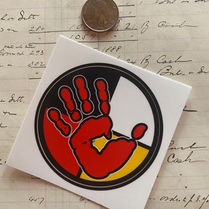 May include: A round sticker with a black outline and a red handprint in the center. The handprint is surrounded by four colored sections: black, red, yellow, and white. The sticker is on a white background.
