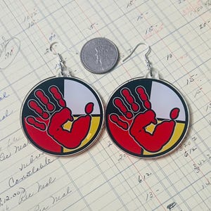 May include: A pair of earrings featuring a red handprint design on a circle with four colored sections: black, white, red, and yellow. The earrings are made of metal and have a silver finish.
