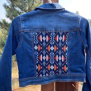 May include: A blue denim jacket with a colorful geometric pattern on the back. The pattern is in shades of red, orange, blue, white, and black.
