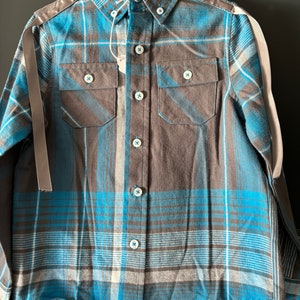 May include: A blue and brown plaid long-sleeve button-down shirt with white accents. The shirt has two chest pockets and a white tag attached to the collar.
