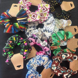 May include: A collection of colorful fabric scrunchies with various patterns, including geometric shapes, floral designs, and stripes. Each scrunchie is attached to a brown cardboard tag. The scrunchies are in shades of blue, pink, green, and black.